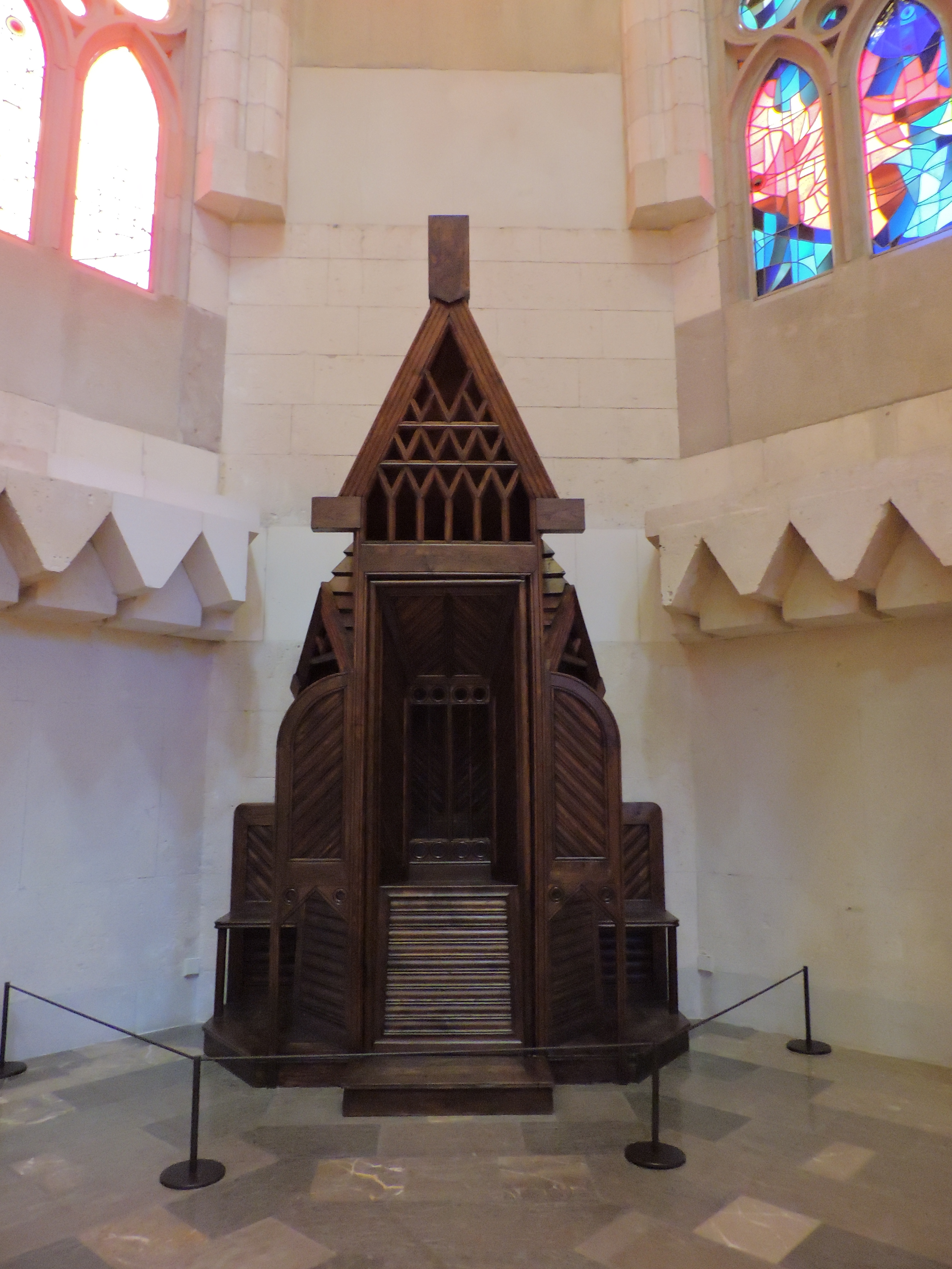 A wooden altar or pulpit located in a church, featuring intricate carvings and a pointed roof, illuminated by stained glass windows.